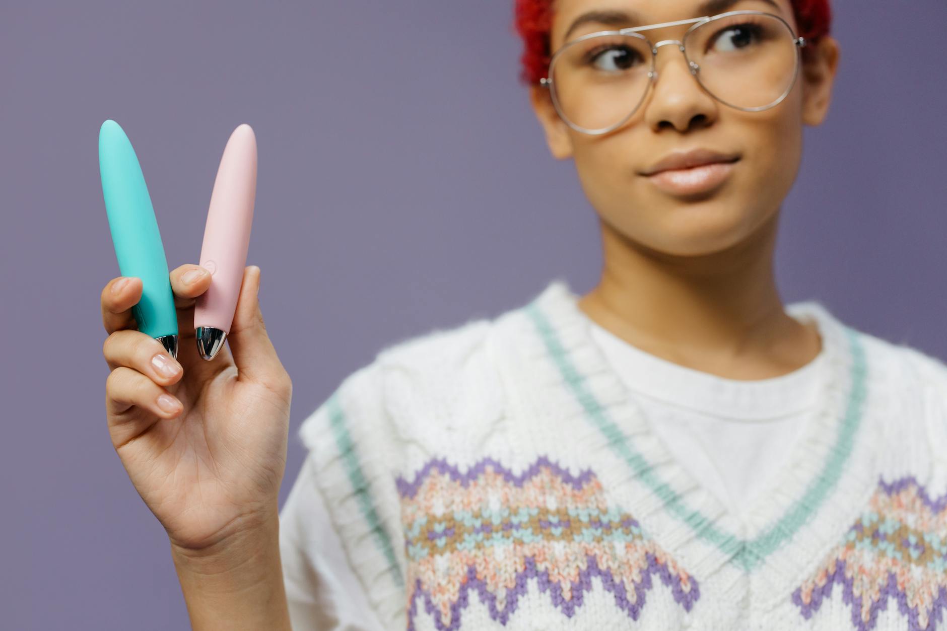 Mulher jovem com óculos segurando dois vibradores coloridos contra um fundo roxo